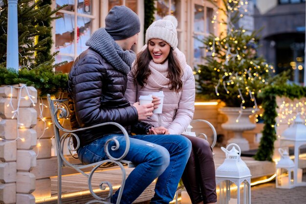 A young romantic couple wearing warm clothes sitting on a bench in evening street decorated with beautiful lights, talking and warming with coffee at Christmas time outdoor