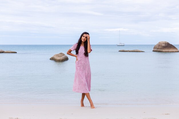 Молодая романтичная женщина с длинными темными волосами в платье на пляже, улыбаясь и смеясь, хорошо проводит время в одиночестве