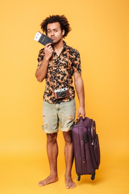 Молодой грустный африканский человек, держащий паспорт и чемодан.