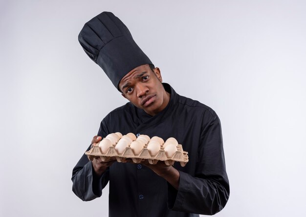 Young sad afro-american cook in chef uniform holds batch of eggs isolated on white wall 