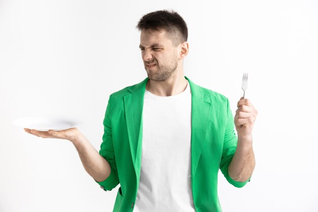 Young sad attractive caucasian guy holding empty dish and fork isolated on grey background. Copy space and mock up. Blank template background. Reject, rejection concept