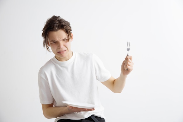 Young sad attractive caucasian guy holding empty dish and fork isolated on grey