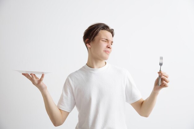 Young sad attractive guy holding empty dish and fork isolated on grey wall.