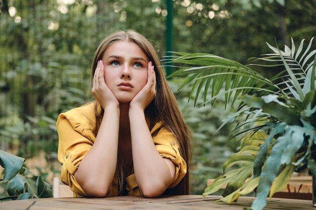 Young sad brown haired woman in yellow shirt leaning head on hands thoughtfully looking up around green leaves in city park