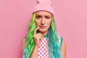 Free photo young sad female model with long dyed hair keeps finger near corner of lips looks displeased has sulking expression wears hat checkered dress isolated on pink