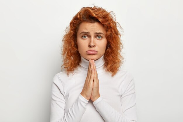 Young sad girl asks for something, keeps hands in praying gesture, pleads for help, purses lower lip, looks with miserable face expression , has foxy curly hair, healthy skin. I am so sorry