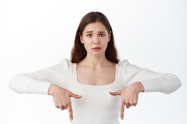 Young sad girl look with anxious and upset face pointing fingers down at something upsetting showing bad news on bottom copy space standing against white background