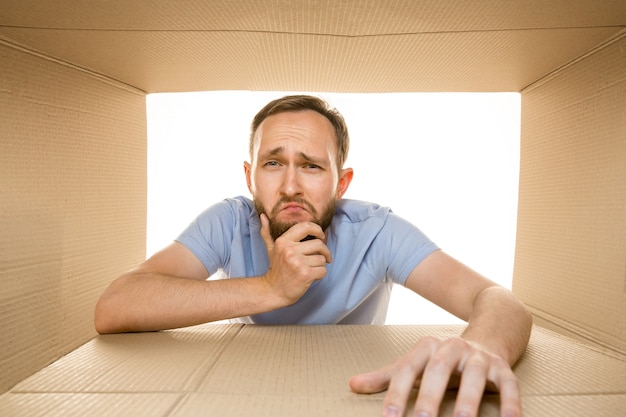 Free photo young sad man opening the biggest postal package isolated on white. dissapointed male model on top of cardboard box looking inside.