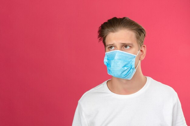 Young sad thoughtful man in medical protective mask looking away and thinking while standing over isolated pink background