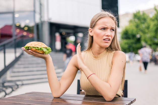 Young sad woman holding burger not satisfied while sitting in outdoors fast food