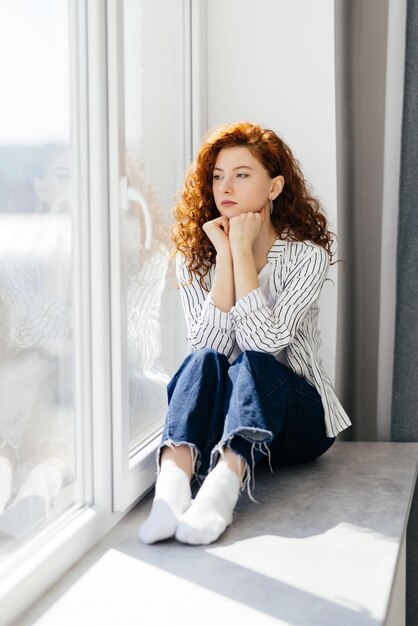 Young sad woman with red hair is sitting on the windowsill