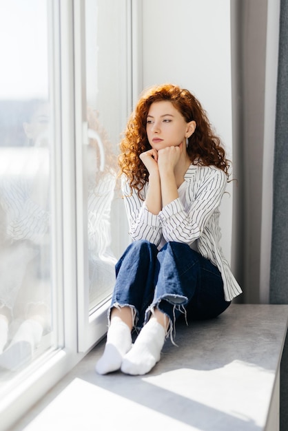 Young sad woman with red hair is sitting on the windowsill