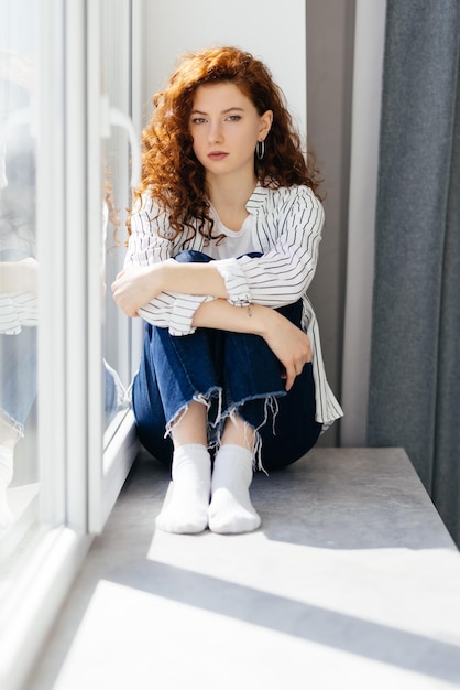 Free photo young sad woman with red hair is sitting on the windowsill