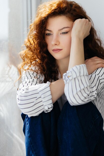 Молодая грустная женщина с рыжими волосами сидит на подоконнике
