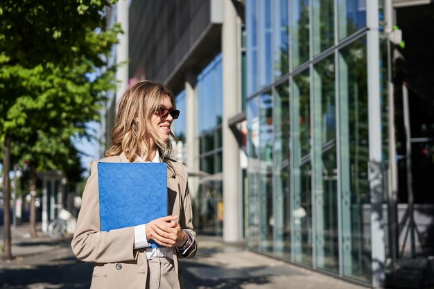 Молодая продавщица собирается работать в солнцезащитных очках и костюме, держа папку с документами, идущую по улице