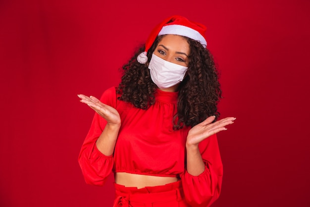 Covid-19 코로나바이러스 바이러스를 입은 크리스마스 모자를 쓴 젊은 산타클로스 여성은 빨간색 배경 스튜디오에 격리된 마스크를 저장했습니다. 새 해 복 많이 받으세요 축 하 휴일 개념입니다.