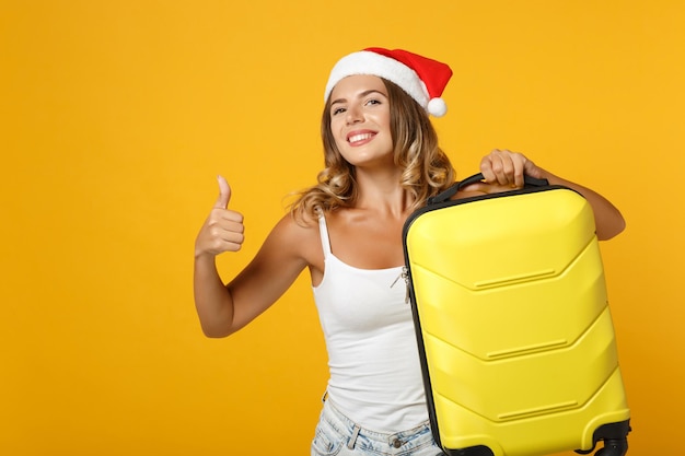 Молодая девушка санта в белой рубашке, рождественская шляпа позирует изолированно на желтом фоне в студии. концепция праздника празднования нового 2020 года. скопируйте пространство для копирования. держа чемодан, показывая большой палец вверх.