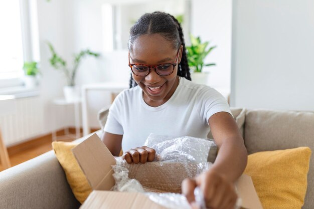 Молодая довольная счастливая африканская девушка женщина леди шопоголик клиент сидит на диване распаковывает коробку доставки посылки онлайн концепция отгрузки покупок