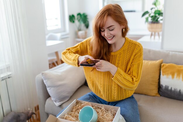 만족한 젊은 여성 쇼핑 중독자 고객은 소파에 앉아 소포 배달 상자 온라인 쇼핑 배송 개념을 풀고 제품 사진을 찍어 소셜 미디어에 게시합니다.
