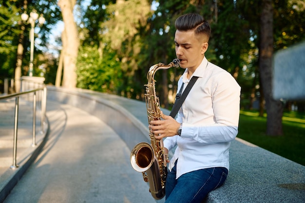 Молодой саксофонист играет на саксофоне в летнем парке. музыкант с саксофоном на открытом воздухе, музыкальное выступление на природе, джазовый исполнитель