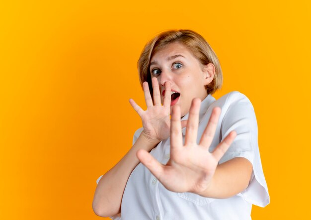 Young scared blonde russian girl defends with hands isolated on orange background with copy space
