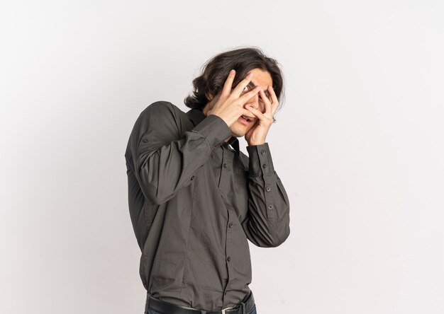 Young scared handsome caucasian man looks through fingers isolated on white background with copy space