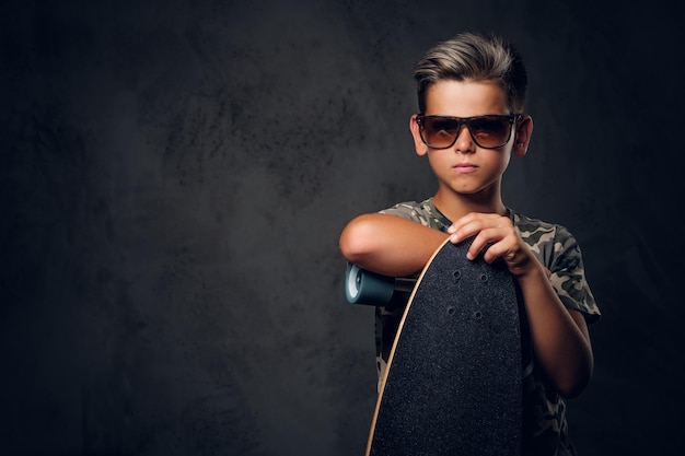 Il giovane scolaro in occhiali da sole sta posando in uno studio fotografico scuro con il suo skateboard.