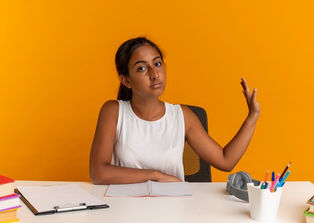 眼鏡をかけている若い笑顔のかわいい白人女子高生は 学校のツールで机に座って時計を保持し コピースペースでオレンジ色のスペースに分離された手を上げます 無料の写真