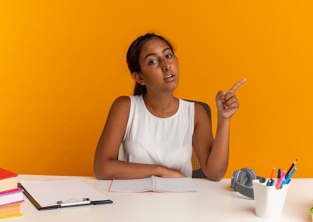 young schoolgirl sitting at desk with school tools points at side 