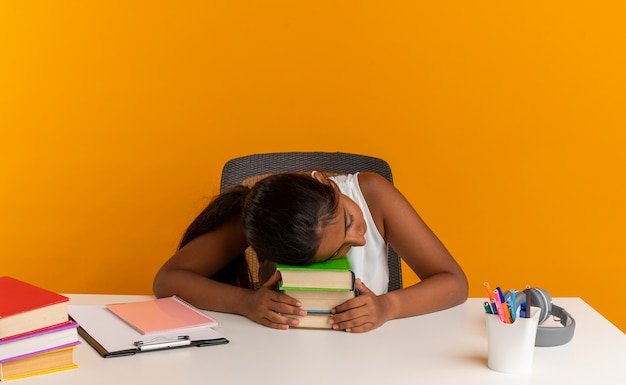 Young schoolgirl sitting at desk with school tools putting head on books isolated on orange wall