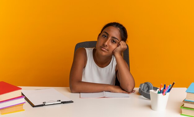 young schoolgirl sitting at desk with school tools putting head on hand 