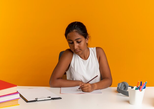 Young schoolgirl sitting at desk with school tools  write something on notebook on orange