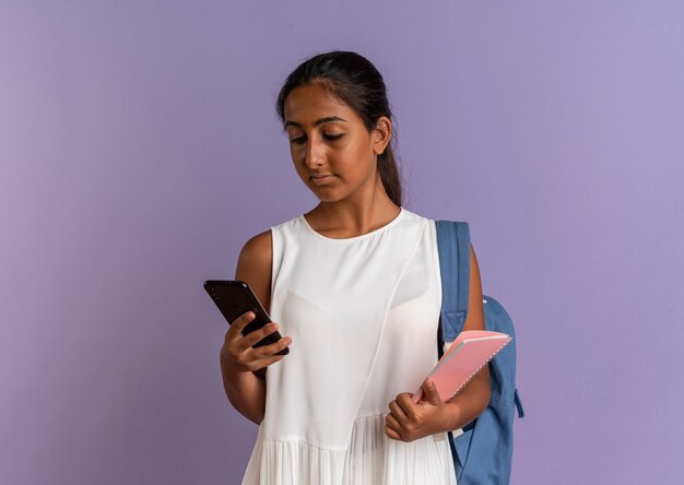 Young schoolgirl wearing back bag holding notebook and looking at phone in her hand on purple