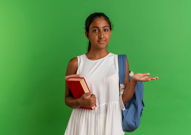 young schoolgirl wearing backpack holding book with notebook and showing with hand at side 