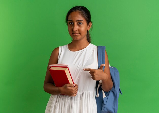 young schoolgirl wearing backpack holding and points at book with notebook 