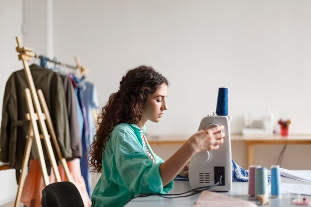 Young seamstress with dark curly hair in colorful shirt dreamily using sewing machine in modern sewing workshop