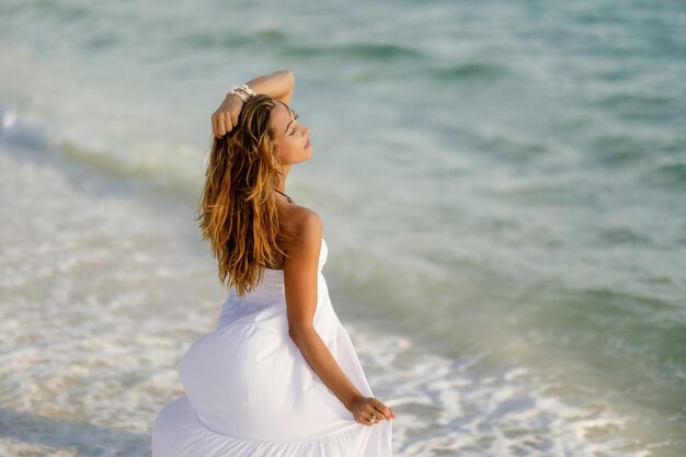 Молодая чувственная женщина в сарафане стоит на берегу с закрытыми глазами и наслаждается летними каникулами