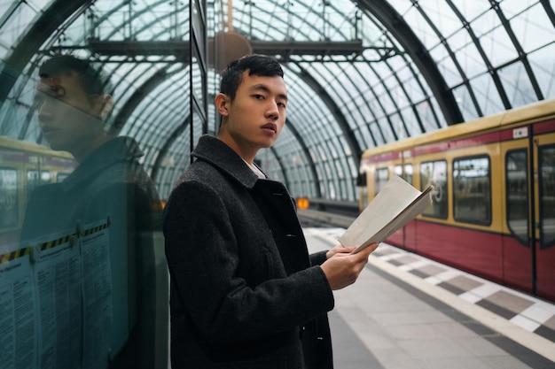 Молодой серьезный азиатский бизнесмен с газетой уверенно смотрит в камеру, ожидая поезда на станции метро