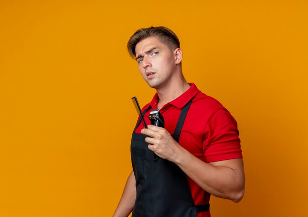 Молодой серьезный светловолосый парикмахер в униформе держит расческу и машинку для стрижки волос, изолированную на оранжевом пространстве с копией пространства