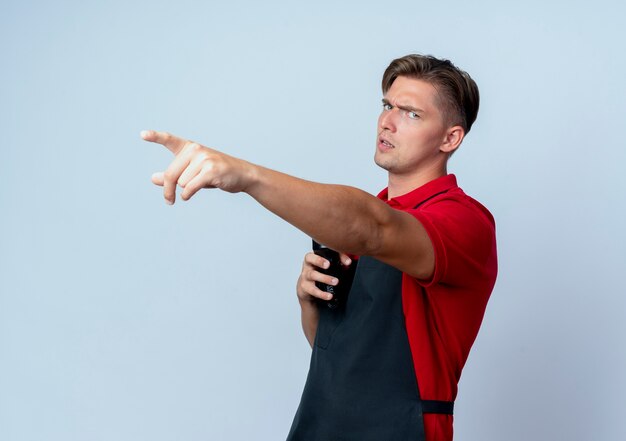 young serious blonde male barber in uniform holds hair clipper pointing at side