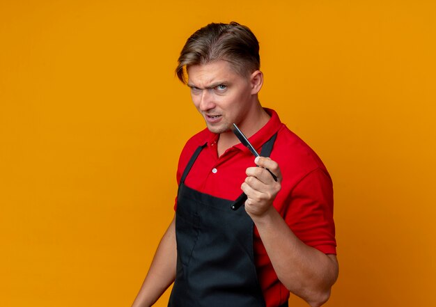 Young serious blonde male barber in uniform holds straight razor isolated on orange space with copy space