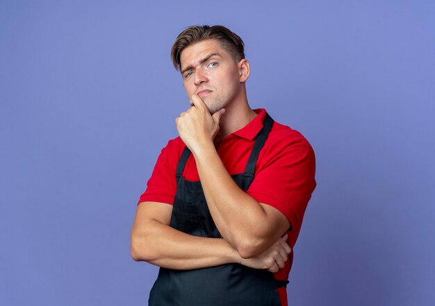 Young serious blonde male barber in uniform puts hand on chin isolated on violet space with copy space