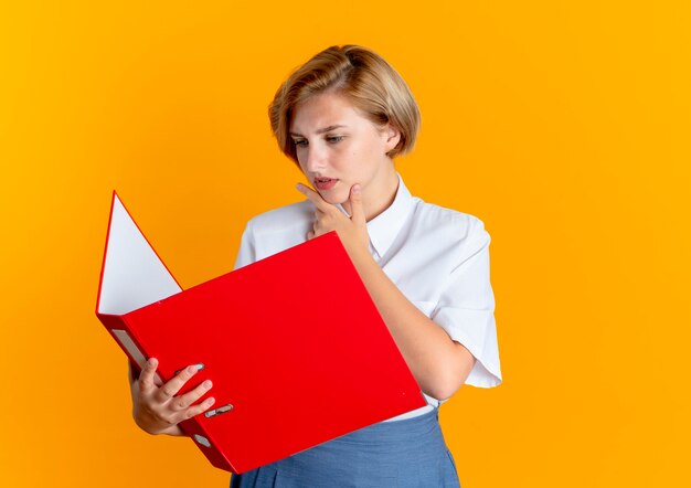 young serious blonde russian girl puts hand on chin looking at file folder isolated on orange background with copy space