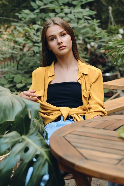 Young serious brown haired teenage girl in yellow shirt and top thoughtfully looking in camera while sitting at wooden table in city park