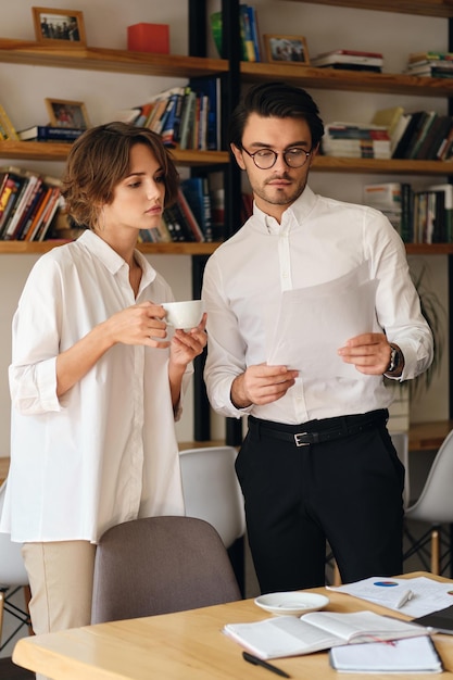 Young serious business colleagues standing near desk with papers while thoughtfully discussing work together in modern office