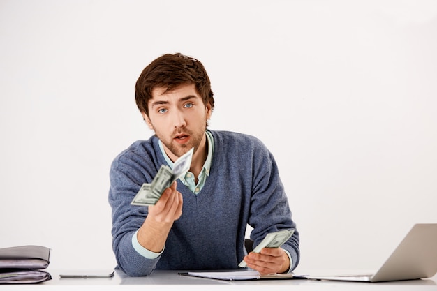 Young serious businessman, counting money sitting office desk with laptop, extend dollars, give half of cash to business partner, making deal