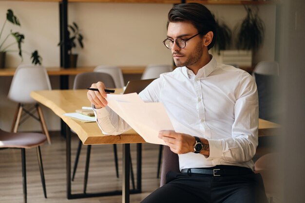 Young serious businessman in eyeglasses thoughtfully working with papers in modern office