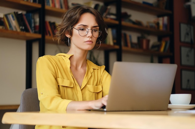 안경을 쓰고 책상에 앉아 있는 진지한 젊은 여성 사업가가 현대 사무실에서 노트북으로 새로운 프로젝트를 신중하게 진행하고 있습니다.