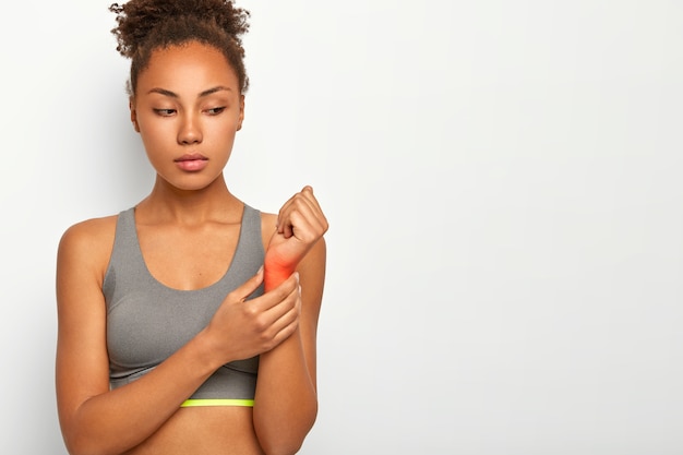 Young serious curly haired woman has injured wrist, raises hand with red focuse showing location of pain, suffers from sprain after workout, wears casual top, poses over white wall with copy space