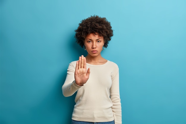 Young serious dark skinned woman shows stop sign prohibition symbol keeps palm forward